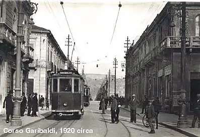 Vintage black and white photograph of Corso Garibaldi in Reggio Calabria, circa 1920, showing a dark tram, pedestrians in early 20th-century clothing, ornate buildings, and a bustling urban street scene.