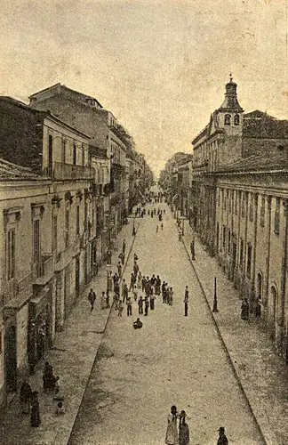 Black and white historical photo of Corso Garibaldi in Reggio Calabria during the 1920s, showing a bustling pedestrian street lined with multi-story buildings, a distant church with a bell tower, and crowds of people in early 20th-century attire.