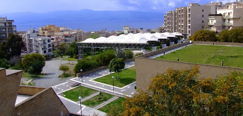 A panoramic view of Parco Caserta Sport Village with lush greenery, swimming pools, and the Straits of Messina in the background.
